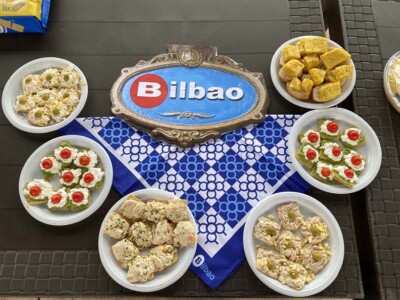 Aste Nagusiaz topera disfrutatzen Una reproducción de las placas de las calles de Bilbao con el nombre de la ciudad, rodeada de platos con pintxos variados