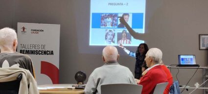 Varios hombres y mujeres mayores participantes en el taller de reminiscencia basada en el fútbol atienden las explicaciones de la dinamizadora del mismo ante una pantalla con imágenes de futbolistas.