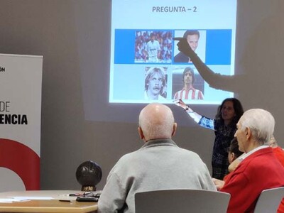 Los recuerdos del fútbol de la Real y el Athletic vuelven a estimular a personas mayores y con daño cerebral Varios hombres y mujeres mayores participantes en el taller de reminiscencia basada en el fútbol atienden las explicaciones de la dinamizadora del mismo ante una pantalla con imágenes de futbolistas.