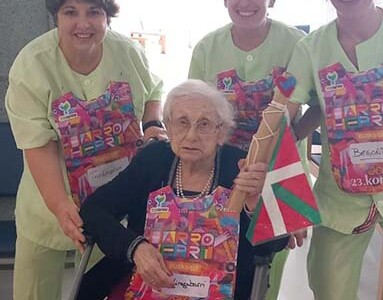El testigo de la Korrika recorre la Residencia Txurdinagabarri Tres mujeres auxiliares de enfermería acompañan a una mujer mayor en silla de ruedas que sostiene el testigo de la Korrika