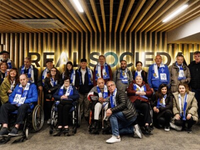 Un amplio grupo de personas posa con bufandas de la Real Sociedad en las instalaciones del estadio Reale Arena