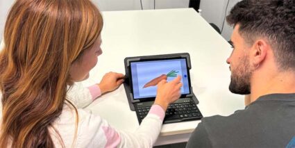 Pacientes de Hermanas Hospitalarias Aita Menni prueban un innovador test para valorar el grado de afasia Una mujer y un hombre jóvenes, de espaldas, ante un pequeño ordenador portátil realizando un test