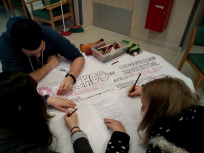 Dos chicas y un chico joven con discapacidad intelectual, sentados en una mesa elaborando un cartel en el que puede leerse 