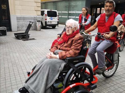 En bici a cualquier edad En primer plano, una mujer de avanzada edad, sentada en una silla de ruedas sobre la plataforma de un triciclo que conduce un voluntario de Cruz Roja en una calle de Bilbao