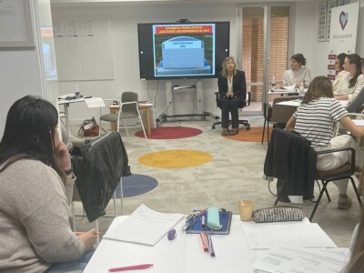 Varias personas atienden las explicaciones de otra, sentada junto a un monitor con una presentación