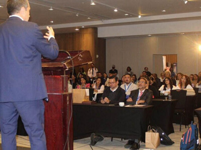 Un hombre vestido con traje azul, de espaldas, pronuncia una conferencia frente a los asistentes en un auditorio