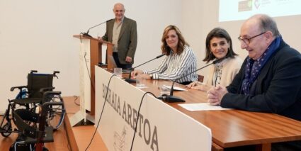 Recogemos donaciones de ayudas técnicas en Bilbao y Durango para su reutilización Dos mujeres junto a un hombre en una mesa presidencial de una sala. Al fondo, un hombre junto a un atril.