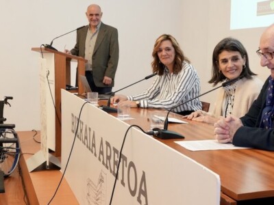 Recogemos donaciones de ayudas técnicas en Bilbao y Durango para su reutilización Dos mujeres junto a un hombre en una mesa presidencial de una sala. Al fondo, un hombre junto a un atril.