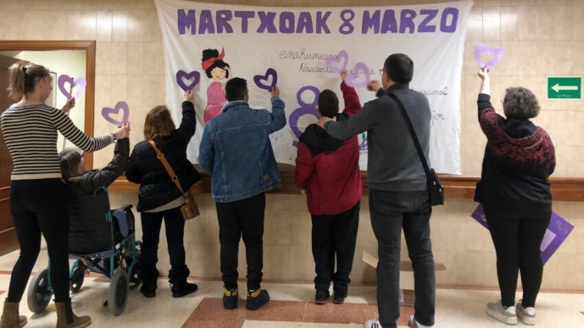 Varias personas jóvenes, de espaldas, delante de un mural sobre el 8M