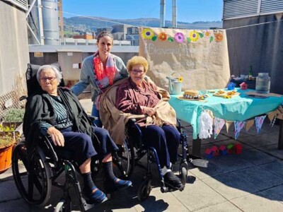 Udaberria etorri dela ospatu dugu Txurdinagabarriko goiko terrazan Dos mujeres mayores en silla de ruedas y una monnitora, junto a una mesa con un picoteo y un cartel alusivo a la primavera