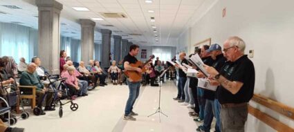 Un coro, junto a un guitarrista, delante de un amplio grupo de personas mayores sentadas