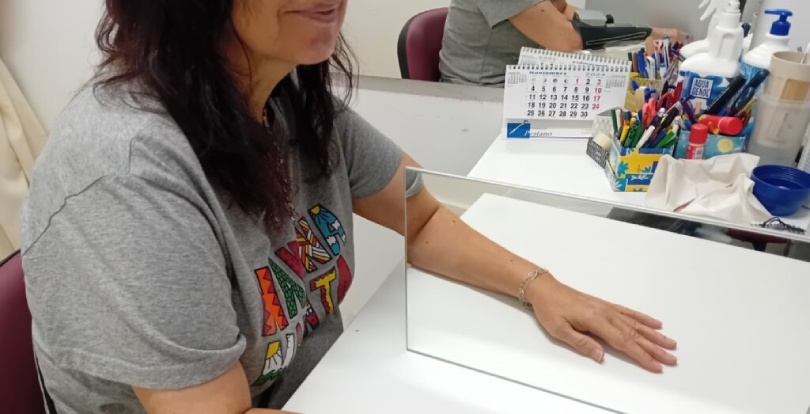 Rehabilitación robótica del miembro superior: innovación funcional en evolución Una mujer, sentada ante una mesa en la que un espejo refleja uno de sus brazos