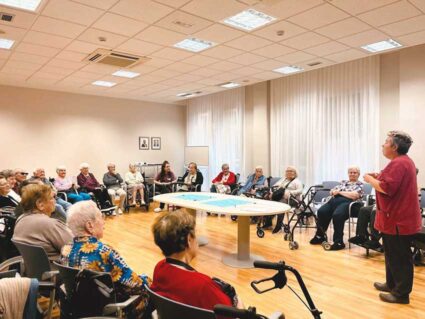 Un grupo amplio de personas mayores, sentadas en círculo en una sala escuchando a una mujer con uniforme rojo de monitora