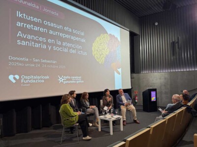 Cinco personas sentdas en un escenario bajo una pantalla con información sobre la jornada Avances en la atención sanitaria y social del ictus